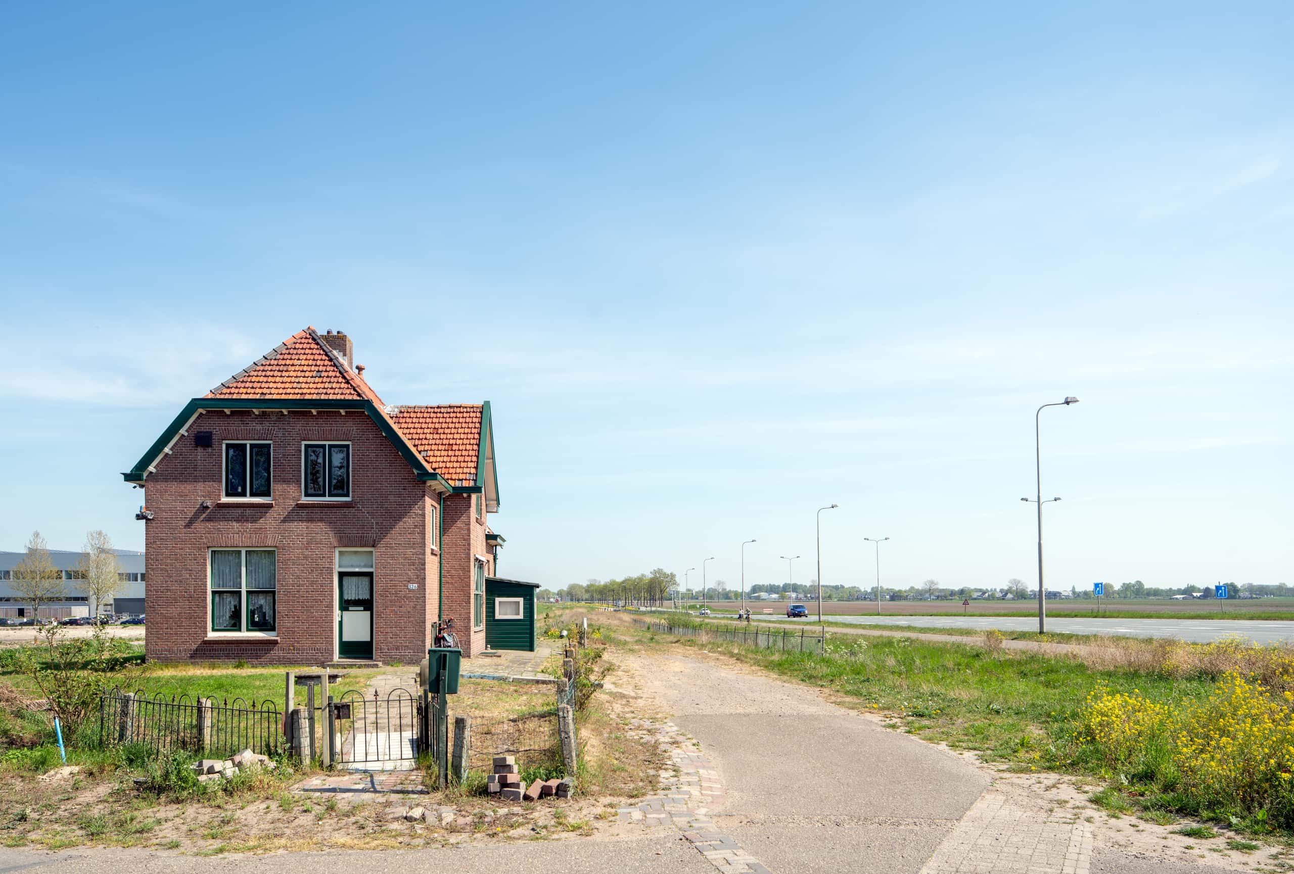 Een vrijstaande woning aan een lege autoweg. De woning heeft een open voortuin met een hekje. Aan weerszijden van de weg gras.