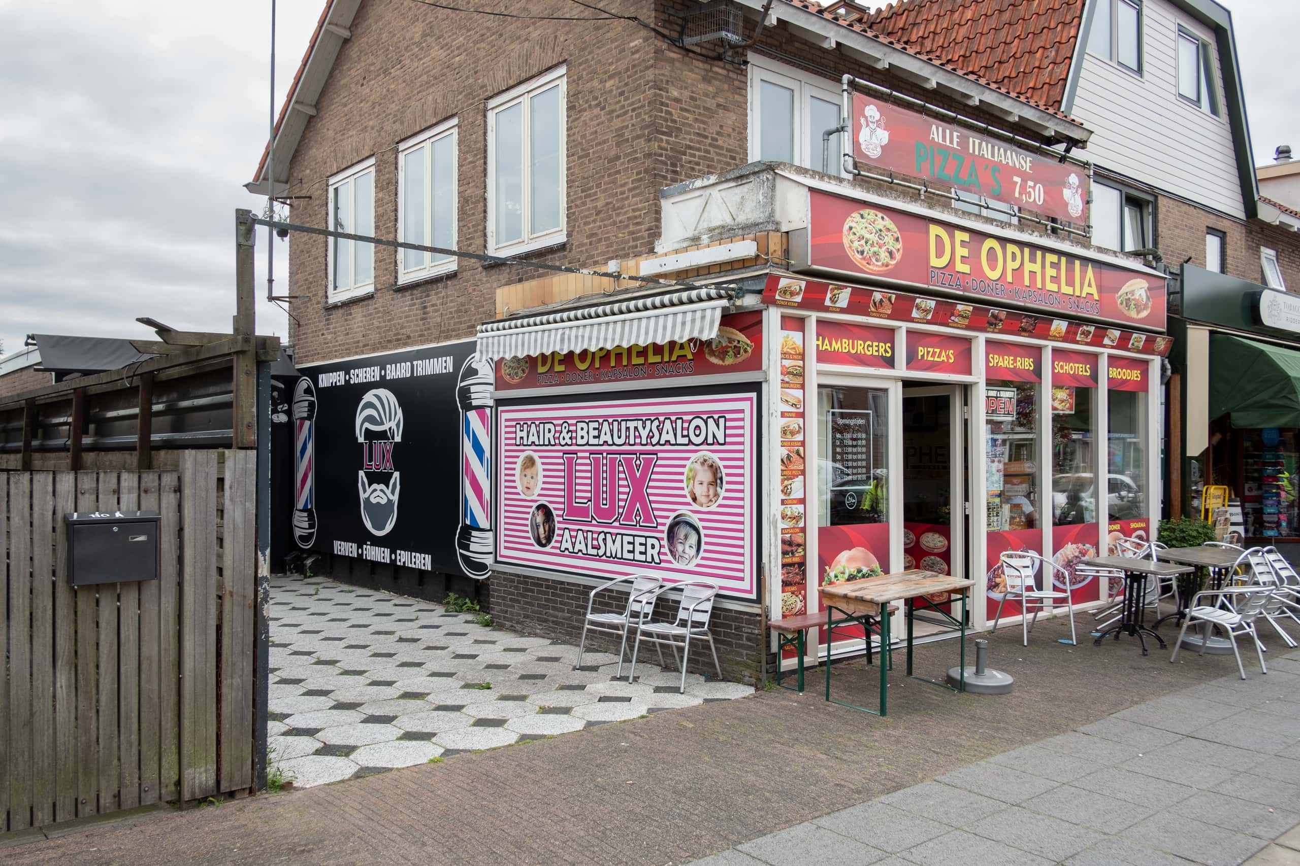 Een gebouw met daarin een snackbar, op de zijkant staat Hair & Beautysalon LUX Aalsmeer op een rozewit gestreepte banner. Aan de voorkant waar de entree is staat een bord met de naam De Ophelia, met daaronder welke gerechten geserveerd worden: hamburgers, pizza's, etc.