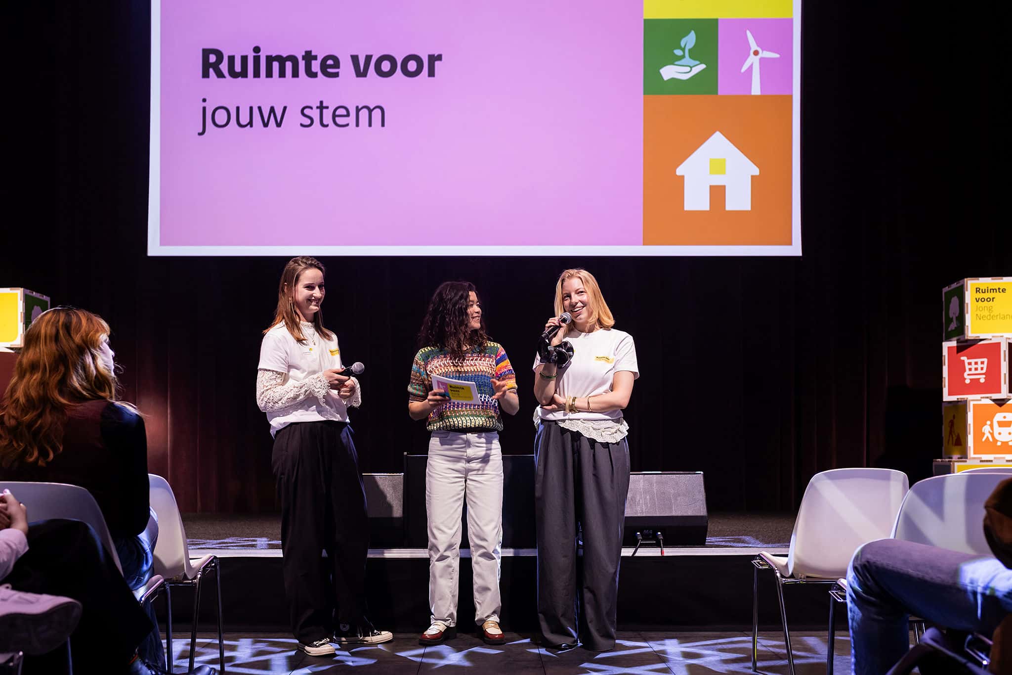 Drie personen op een podium met microfoons; op het scherm staat ‘Ruimte voor jouw stem’ met pictogrammen.