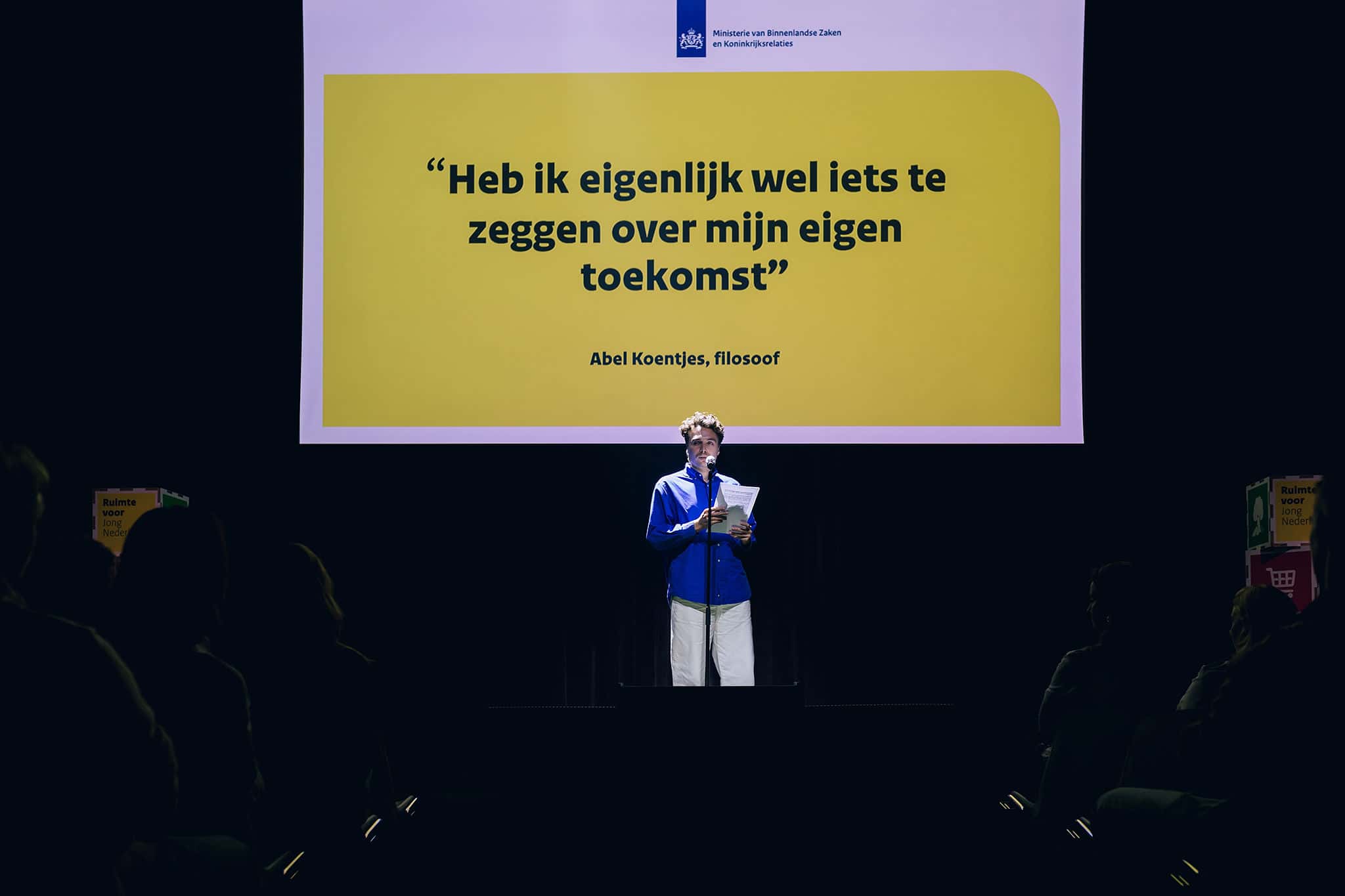 Abel Koentjes leest voor op een podium; op groot scherm staat de quote ‘Heb ik eigenlijk wel iets te zeggen over mijn eigen toekomst’.