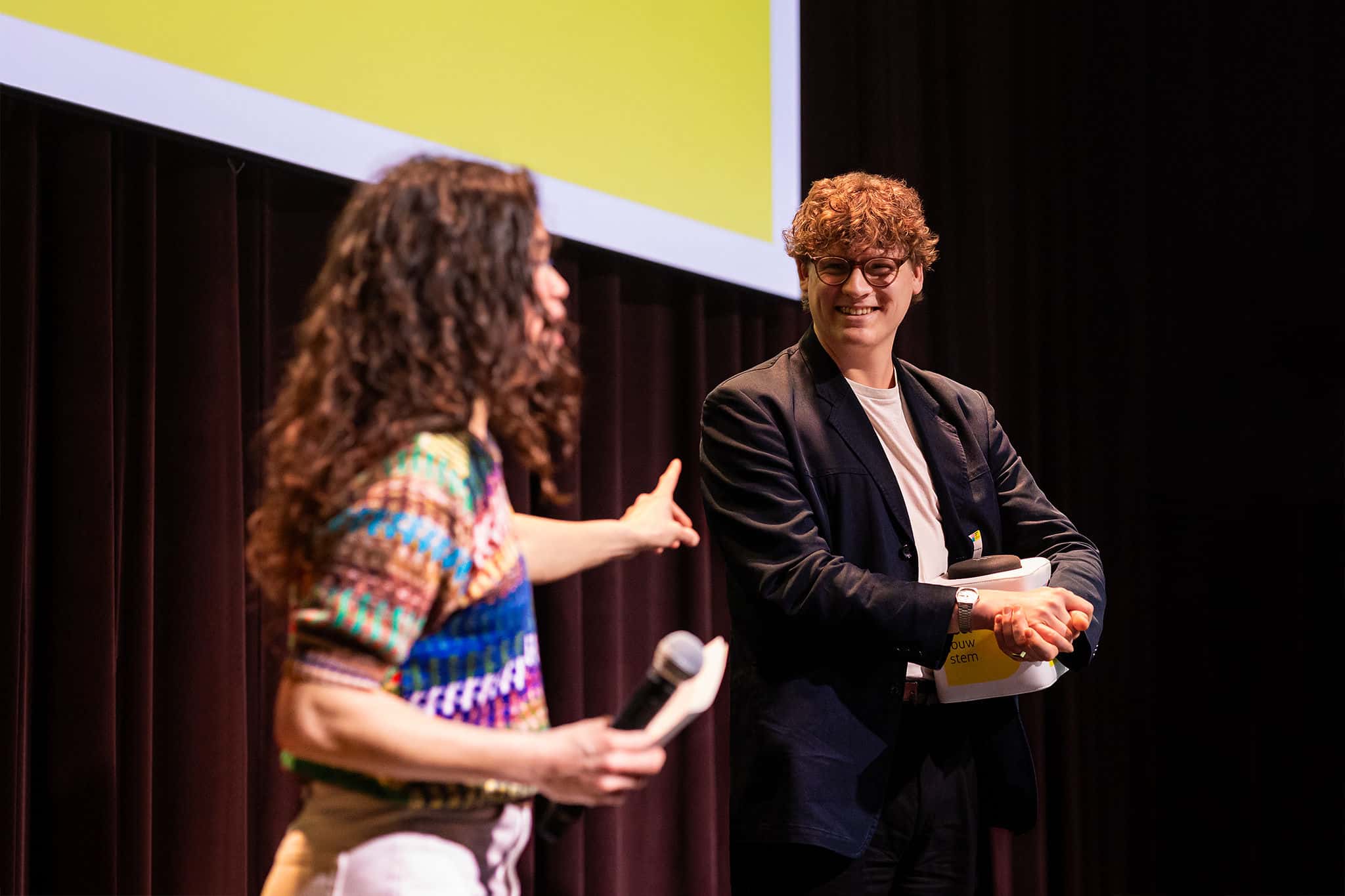 Twee personen op een podium; één stelt een vraag met microfoon, de ander luistert met gevouwen armen voor een groot projectiescherm.
