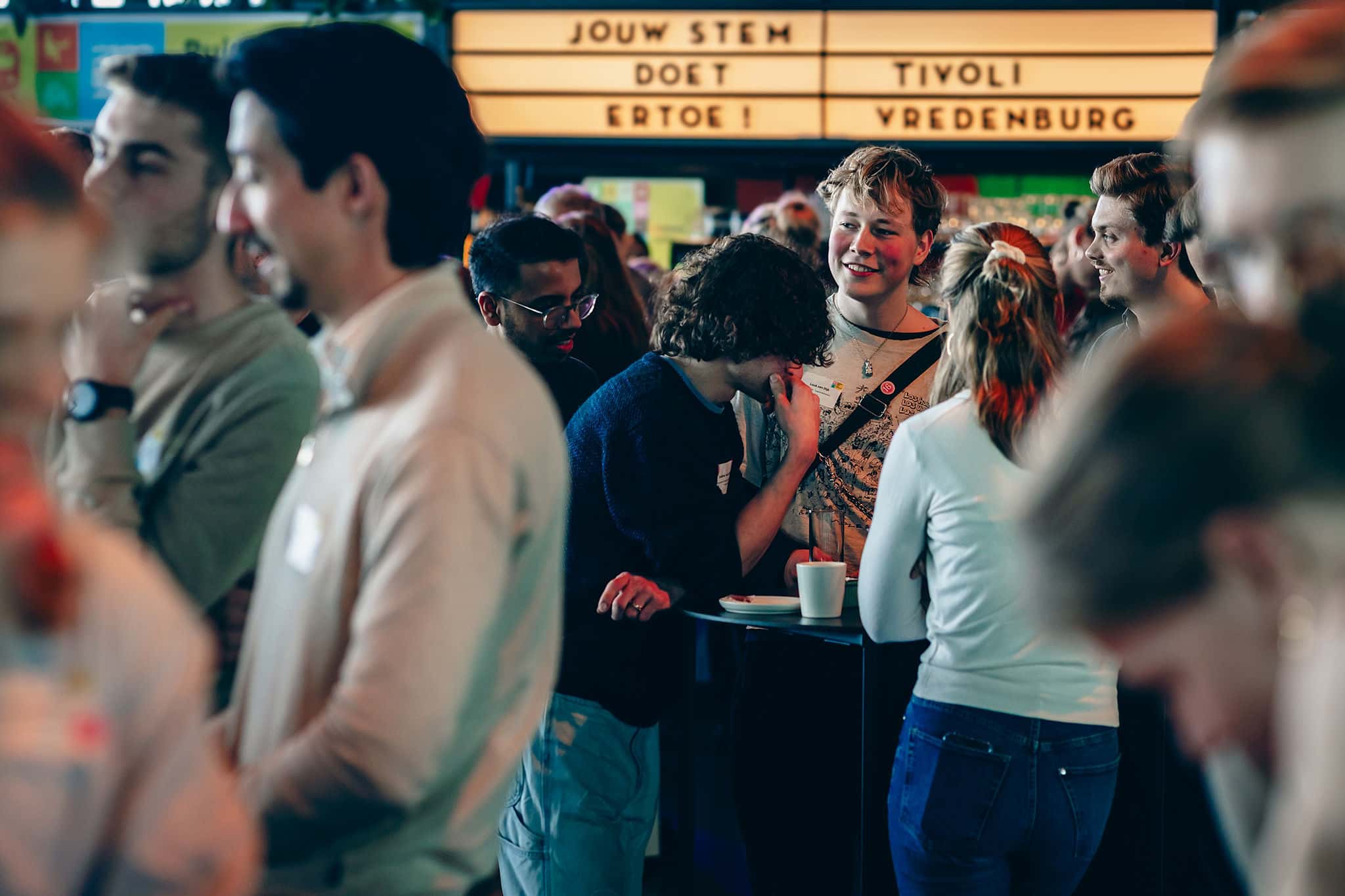 Groep mensen in gesprek bij statafels; op de achtergrond een bord met de tekst ‘Jouw stem doet ertoe’ en ‘TivoliVredenburg’.