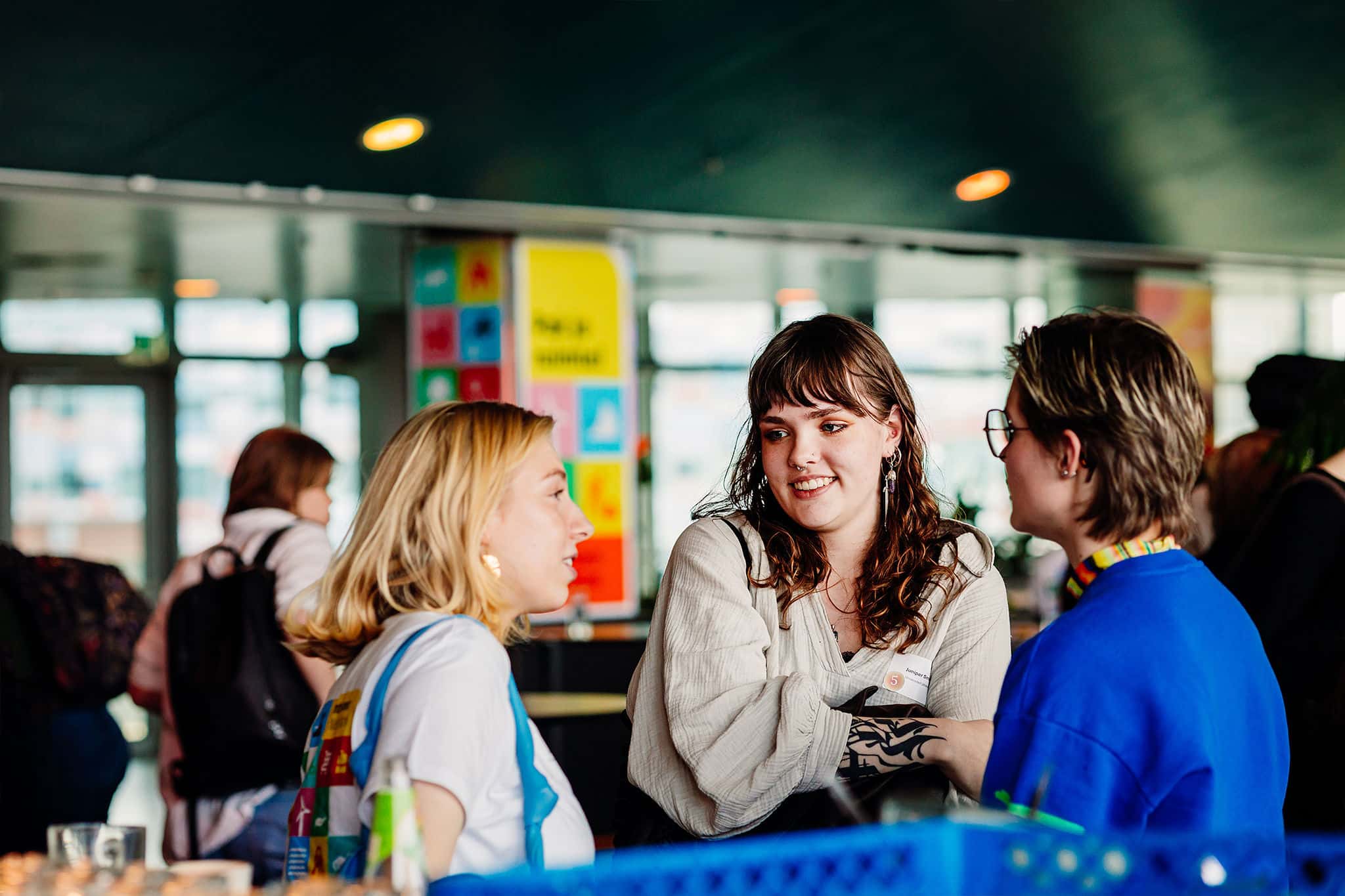 Drie personen staan met elkaar in gesprek in een publieke ruimte; op de achtergrond hangen kleurrijke posters en borden.