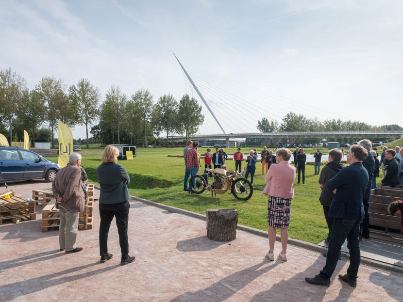 Mensen staan op een grasveld en deels op een parkeerplaats, een fiets staat in het midden en op de achtergrond een brug
