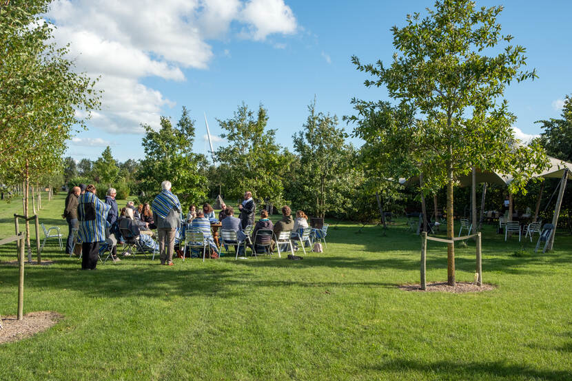 Mensen zitten op een stoel in een cirkel op een grasveld, een aantal mensen staan erachter, rechts staat een tent