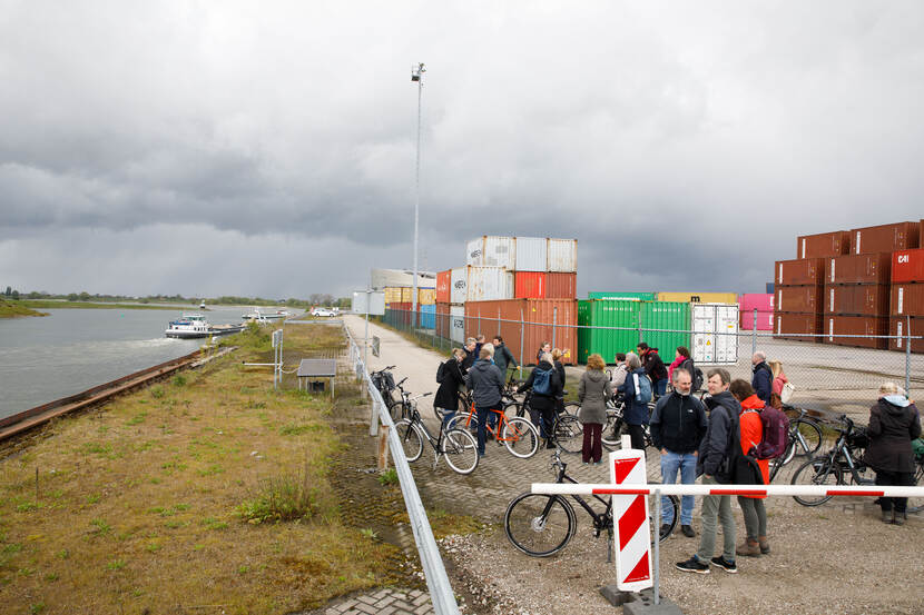 Deelnemers staan met hun fiets aan de hand naar de rivier links te kijken, rechts staan containers