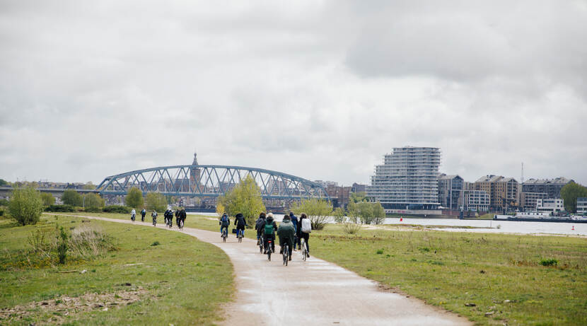 Deelnemers fietsen door groen gebied richting brug en stad
