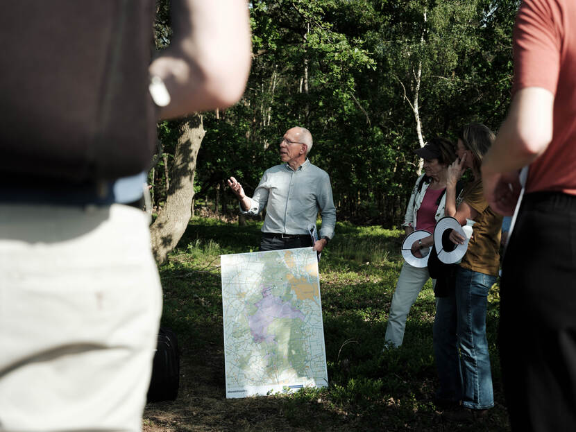 Een man op middelbare vertelt iets aan een groep mensen om hem heen in het bos. Voor zijn voeten heeft hij een grote kaart vast