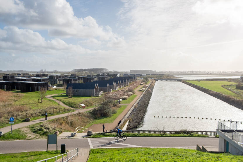 Links veel vakantiewoningen, in het midden een grote strook water, rechts landbouwgrond en op de voorgrond een geasfalteerde weg met een fietser
