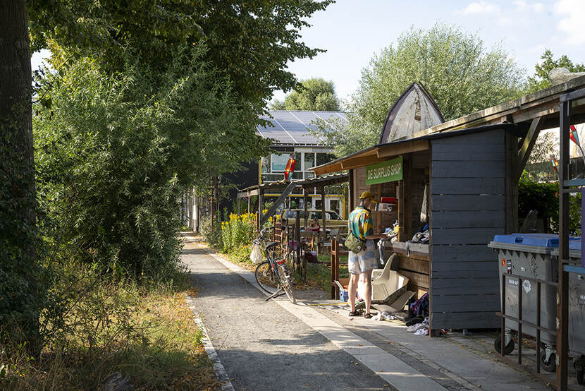 Een man staat langs een fietspad bij een kraampje. Boven het kraampje hangt een banner met daarop 'De surplus shop'. Aan de andere kant van het fietspad is veel groen: bomen, gras en struiken.