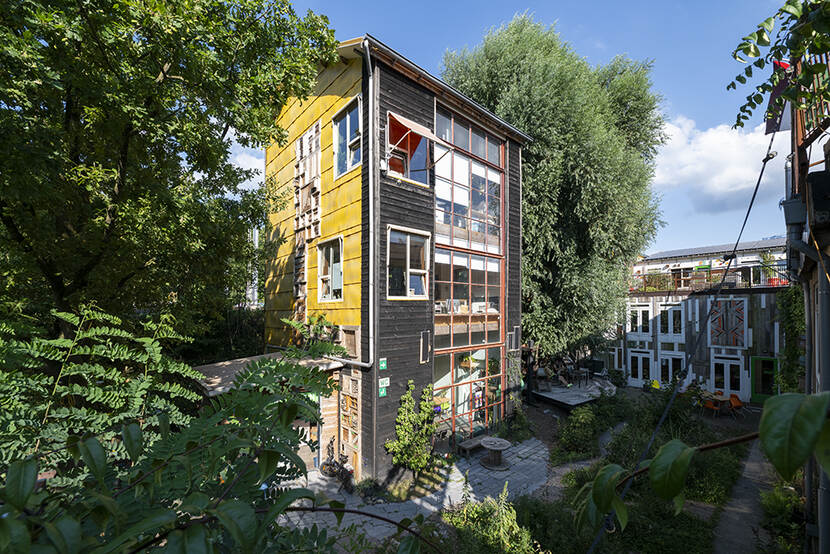 Tussen bomen in staat een smal gebouw van drie verdiepingen. Het is opgebouwd uit meerdere materialen.