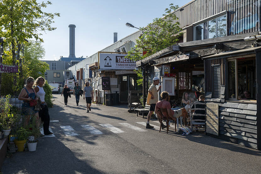 Twee vrouwen lopen over een zebrapad, aan de andere kant van de straat zitten mensen op stoeltjes. Op de achtergrond is de oude fabriek te zien met de schoorstenen. Het zonnetje schijnt.