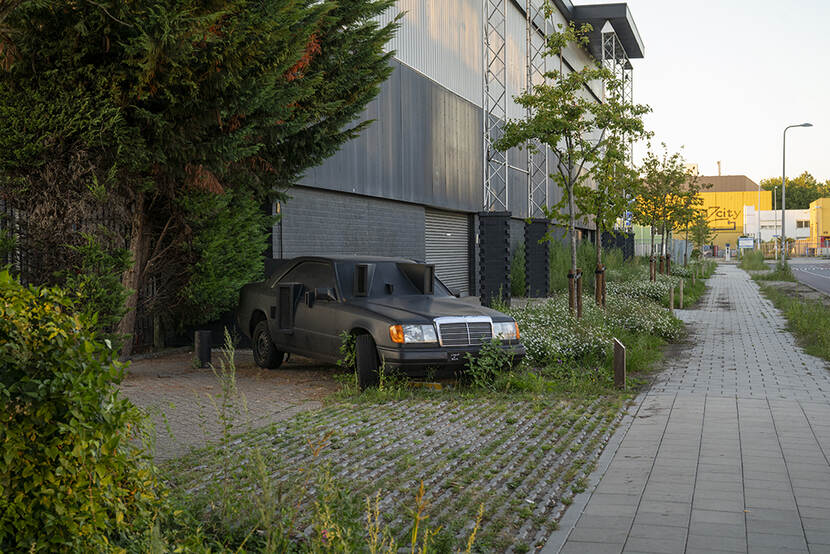 Een zwarte auto staat geparkeerd langs een gebouw. Het is duidelijk dat de auto niet meer in gebruik is, het lijkt door de toegevoegde vormen op de vooruit meer op een kunstinstallatie.