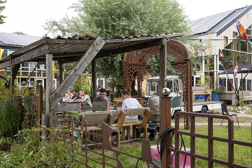 Een vol terras waar mensen aan het werk zijn onder een overkapping. Het terras staat naast een pand met zonnepanelen op het dak.